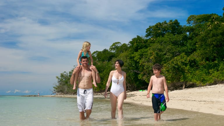 Family walking on the beach in the water with a girl sitting on her father's shoulders.