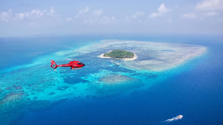 A red helicopter flying over Green Island with a boat sailing below.